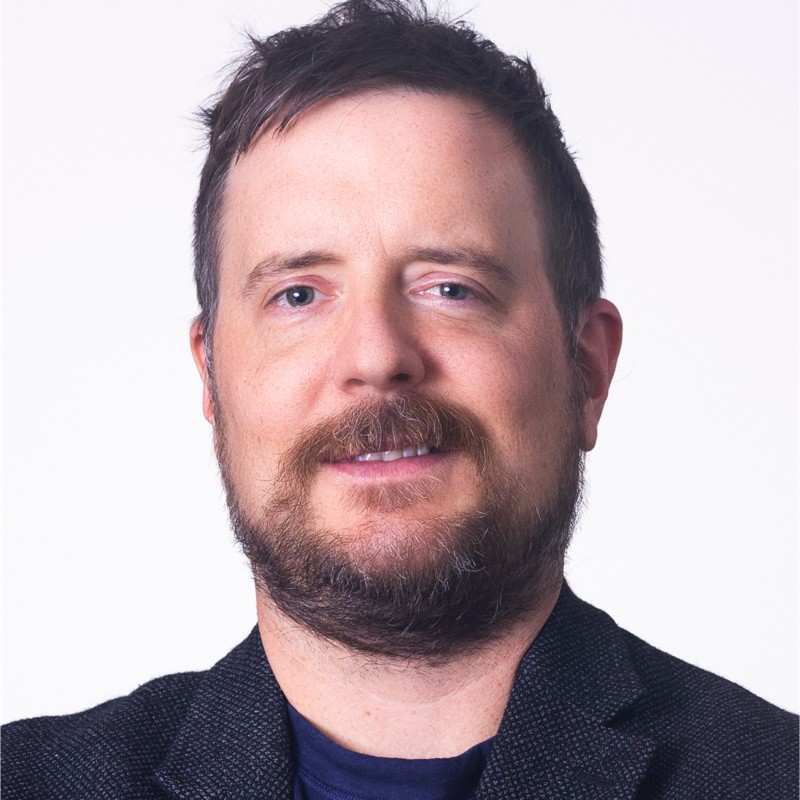 The coach, a man with short dark hair and a beard, in a dark blazer, smiling at the camera
